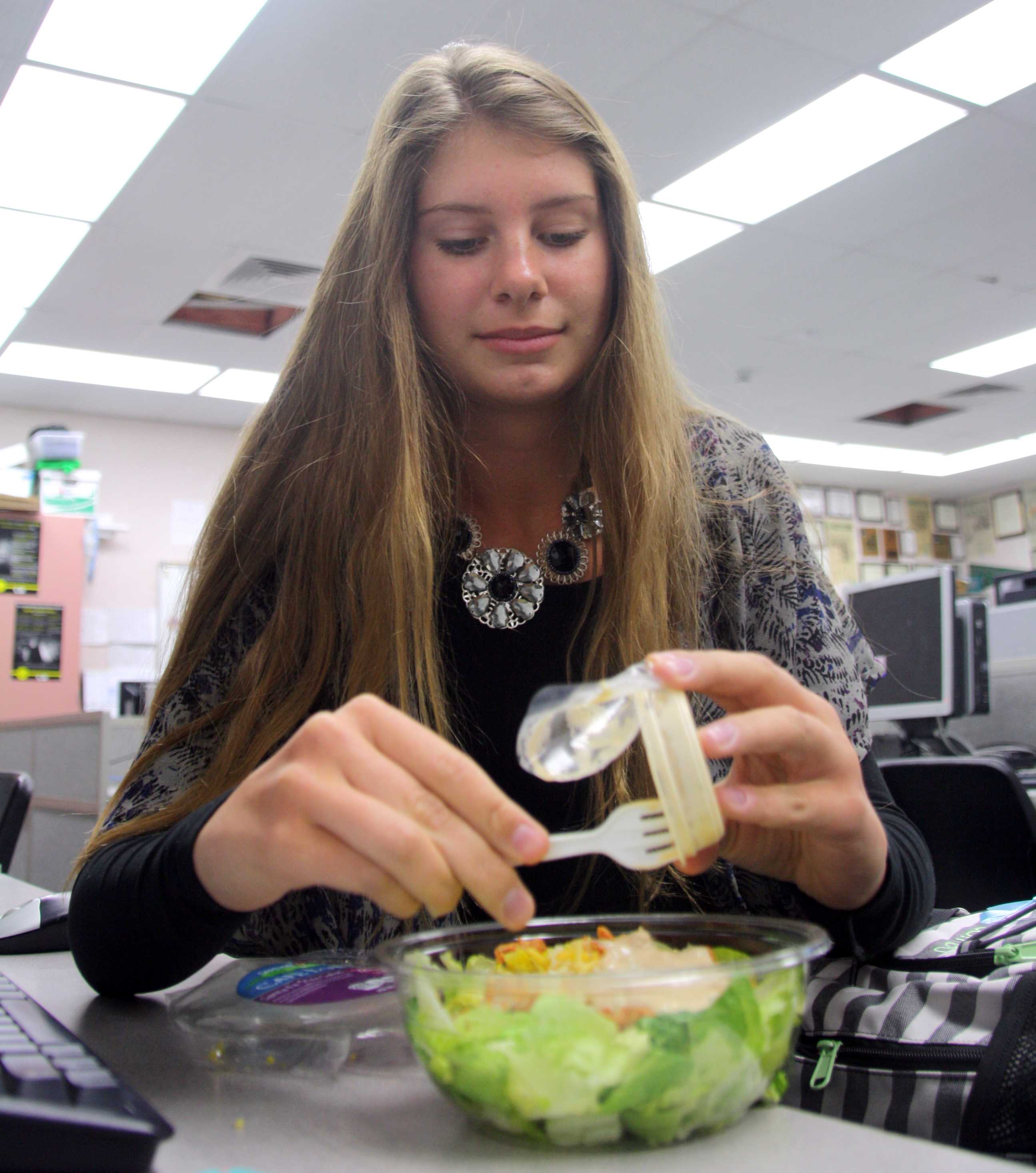 Behind a salad, junior Frances Strnad prepares her Cesar salad on April 16 during lunch. The salad came in package that contained cheese, croutons, and chicken. Not only did she bring a salad, she also brought water and apple slices to accommodate her healthy living style. "Healthy eating is a priority in my household and I really enjoy that. I feel more energized and focused at school. Healthy eating prevents me from becoming lazy and tired," Strnad said. Photo by Ambreen Siddiqui